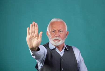 Portrait of mature man with white hair and beard showing stop sign, hand gesture, against blue background in studioの写真素材