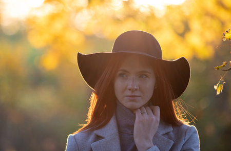 Portrait of pretty redhead girl with hat and in coat walking in forest at sunset in autumnの写真素材