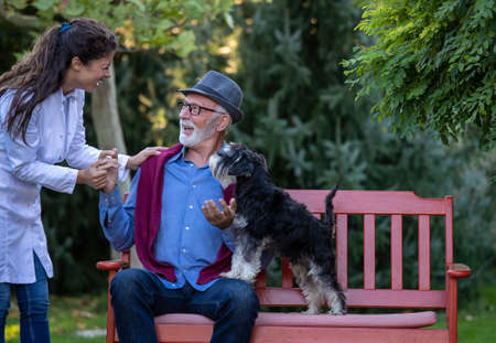 Elderly man with dog sitting in park while pretty nurse in white coat holding hand on his shoulder,...