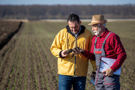 Two farmers checking soil quality in field, senior man holding notepad, while younger carrying lump...