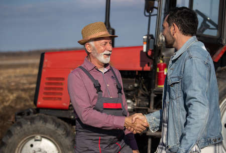 Two farmers senior worker and younger engineer shaking hands in front of tractor in field in autumn timeの写真素材