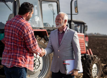 Senior sales representative and mature farmer shaking hands infield in front of tractorの写真素材