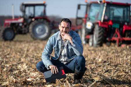 Mature adult farmer squatting in field, thinking and making plans while tractors working in backgroundの写真素材