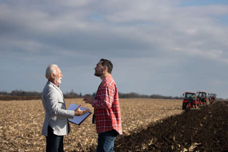 Experienced business man  talking to younger farmer in field in front of tractors in autumnの写真素材