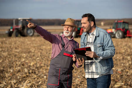 Two farmers standing in field and looking far away. Tractors working in backgroundの写真素材
