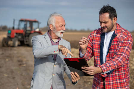 Senior business man showing mature adult farmer where to sign contract in field in front of tractorの写真素材