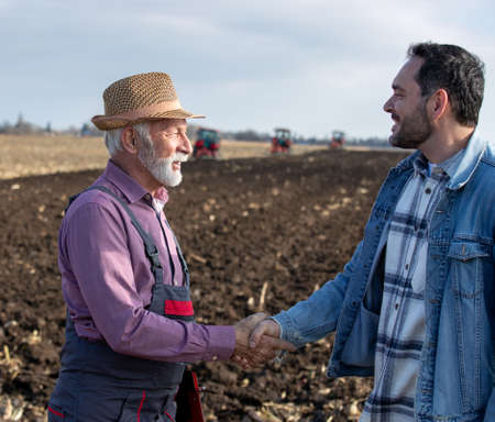 Two farmers senior worker and younger engineer shaking hands in front of tractors in field in autumn timeの写真素材
