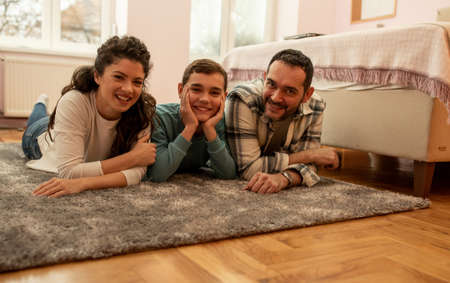 Portrait of happy family of three, father, mother and son lying on floor beside bed in sleeping bedroomの写真素材