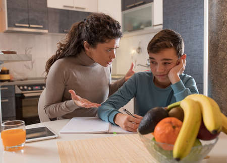 Angry mother yelling on son while doing homework at dining table at home