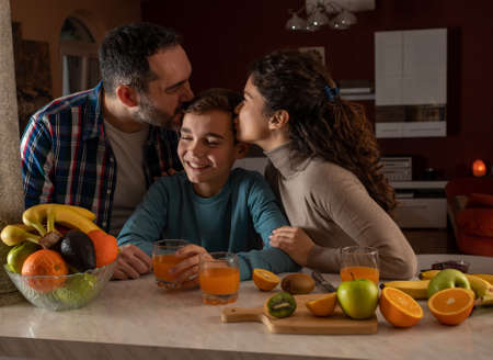Happy family having tender moment at home, mother and father kissing their boy, while preparing juice from fresh fruits on kitchen countertopの写真素材
