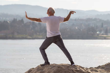 Rear view of senior man practicing tai chi chuan and qigong on sandy river beach, relaxing, meditating in natureの写真素材