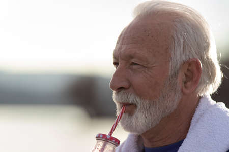 Senior man with beard after workout drinking fresh juice from bottle with strawの写真素材