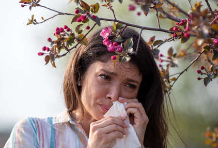 Afraid young woman wiping nose with napkin in front of blooming tree. Spring allergy attack concept