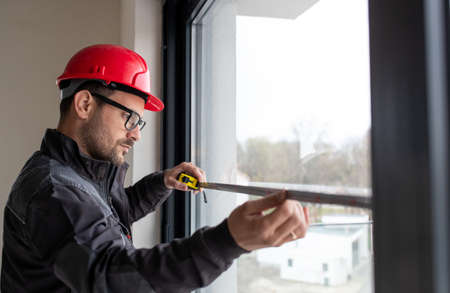 Male worker wearing helmet measuring window with measuring tape for installing blinds indoors.の写真素材