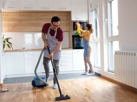 Couple doing their chores together while woman wipes the dust from and man vacuuming floor with...