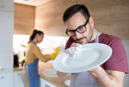 Man with apron in the kitchen polishes plate with a cloth while woman in background wash the dishes.