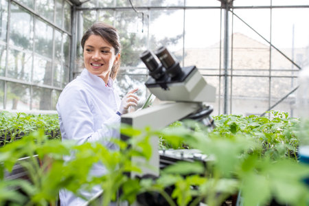 Young pretty woman agronomist in white coat working on tablet and microscope in greenhouse. Plant care and protection conceptの写真素材