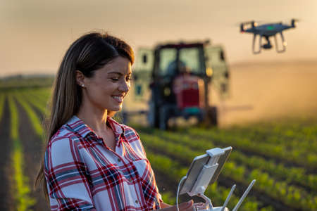 Pretty young farmer woman driving drone with remote control in front of tractor in soybean field in...