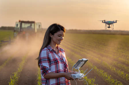 Pretty young farmer woman driving drone with remote control in front of tractor in soybean field in...