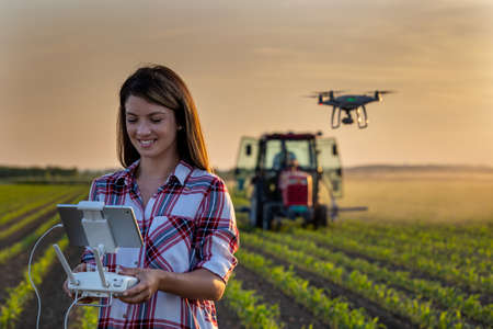 Pretty young farmer woman driving drone with remote control in front of tractor in soybean field in...