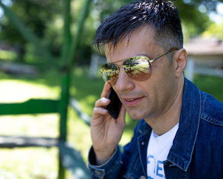 Handsome man in jeans jacket wearing sunglasses in park and talking on mobile phone
