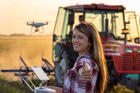 Satisfied young farmer woman driving drone and showing thumb up in field in spring in front of...