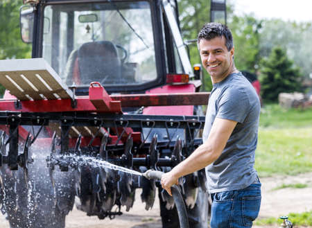 Handsome satisfied man cleaning tractor and equipment with water stream from hose and showing thumb up on farmの写真素材