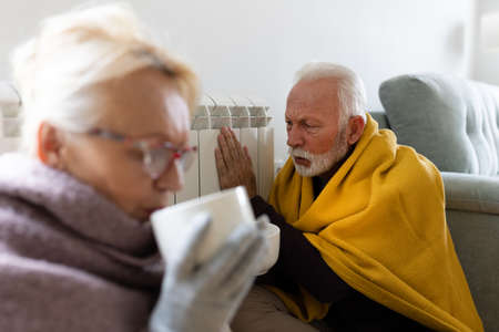 Senior couple covered with blanket sitting beside heater in living room and trying to warm in winter season and energy crisisの写真素材