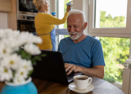 Senior man working on laptop at dining table at home while woman cleaning windows in background