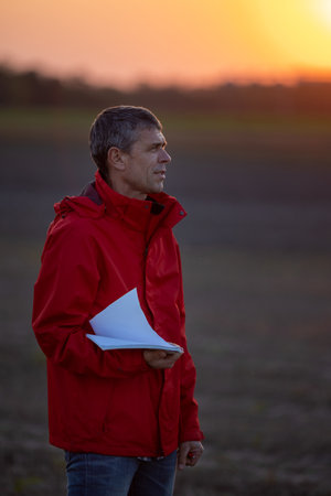 Mature engineer standing in field at sunset in autumn and supervising land