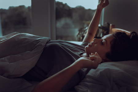 Young pretty woman waking up in bed in early morning with street light coming from windows