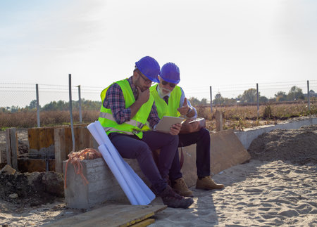 Two engineers with helmets and vests looking at tablet and talking about project at building site