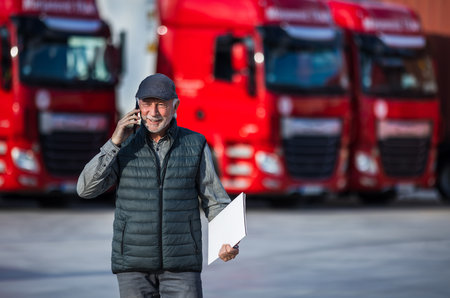 Senior man holding notebook and talking on mobile in front of parked trucks. Carco business conceptの写真素材