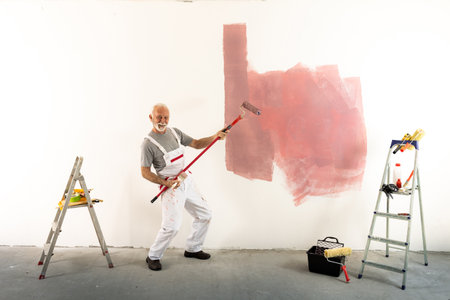 Senior worker in overalls having fun during painting wall, playing guitar on stick of rollの写真素材
