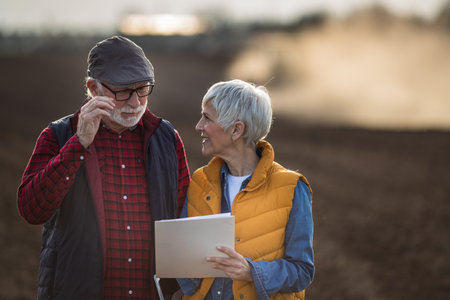 Mature farmers man and woman standing in field and writing notes while tractor plowing in backgroundの写真素材