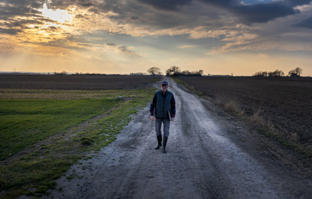 Senior farmer holding laptop in hand and walking on dirty road between two agricultural fields in winter timeの写真素材