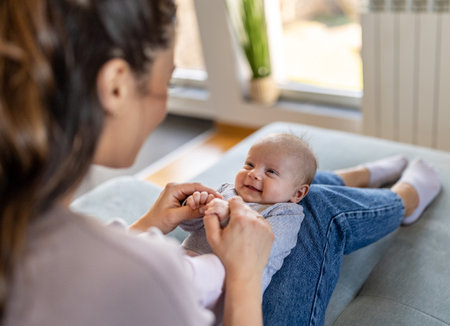 Young mother sitting on sofa at home and holding cute smiling newborn baby on thighs and playing. Mothernity love, care and joy conceptの写真素材