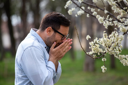 Handsome man sneezing beside blooming tree in spring outdoors. Allergy pollen symptoms conceptの写真素材