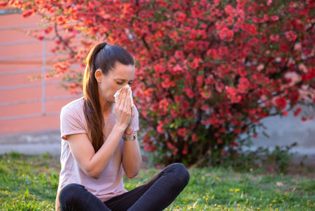 Pretty young woman having alergy symptoms from blooming tree pollen in spring, blowing noseの写真素材