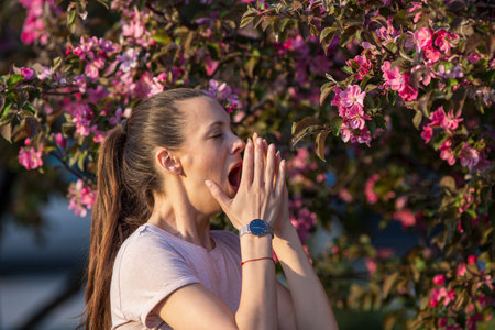 Pretty young woman having alergy symptoms from blooming tree pollen in spring, sneezingの写真素材