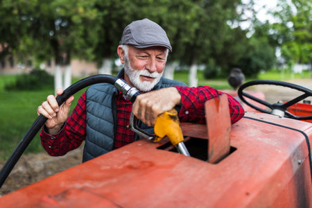 Senior farmer holding fuel pistol and refilling reservoir of tractor on private gas station on farmの写真素材