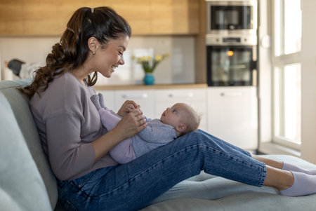 Young mother sitting on sofa at home and holding cute smiling newborn baby on thighs and playing....