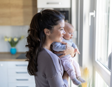 Smiling pretty mum holding infant in arms and enjoying tender moment at home beside window. Mothernity love, care and joy conceptの写真素材