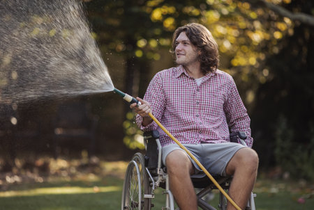 Young smiling man in wheelchair watering plants, splashing from hose in garden