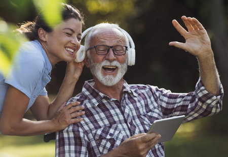 Senior man and young woman nurse listening music from tablet in park on bench and singingの写真素材