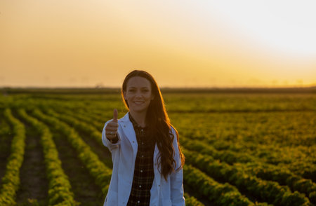 Portrait of young woman agronomist in white coat showing thumb up in soybean field in summer timeの写真素材