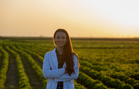 Portrait of young woman agronomist in white coat with crosses arms standing in soybean field during summer, at sunsetの写真素材