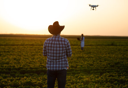 Farmer man and woman standing in field at sunset and driving drone above farmlandの写真素材