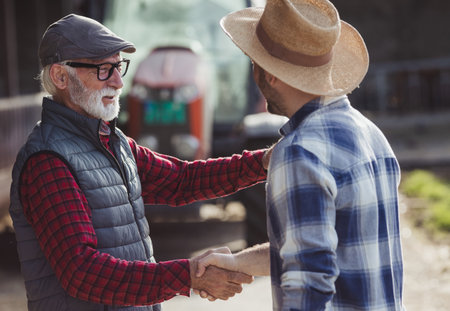 Two farmers young and mature shaking hands in front of tractor on cow farm as sign of deal.の写真素材