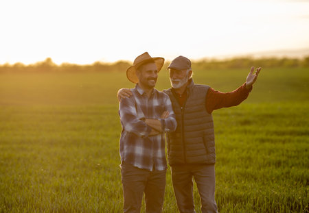 Satisfied senior farmer hugging younger man in wheat field in spring time at sunsetの写真素材